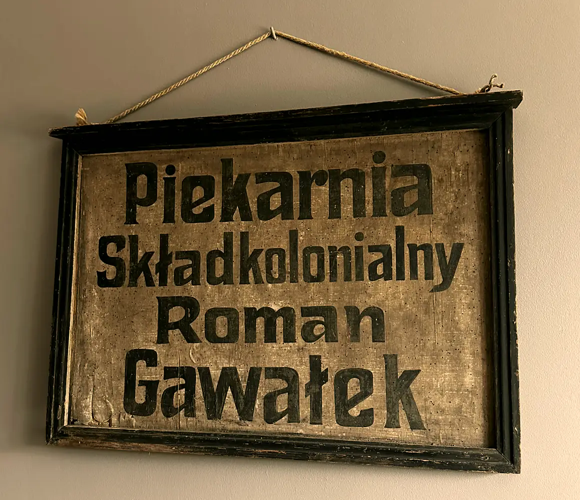Drewniana deska w czarnej, spękanej ramie, zawieszona na sznurku na ścianie; ręcznie malowany napis „Piekarnia Skład kolonialny Roman Gawałek”